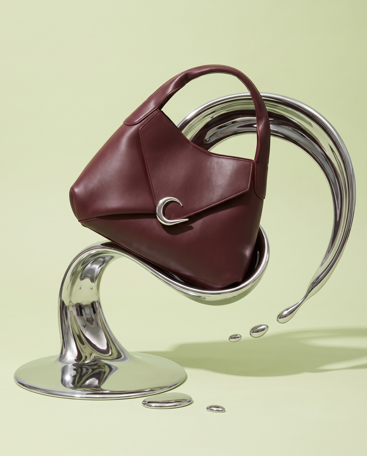 Burgundy leather handbag suspended in a sculptural chrome form against pale green
