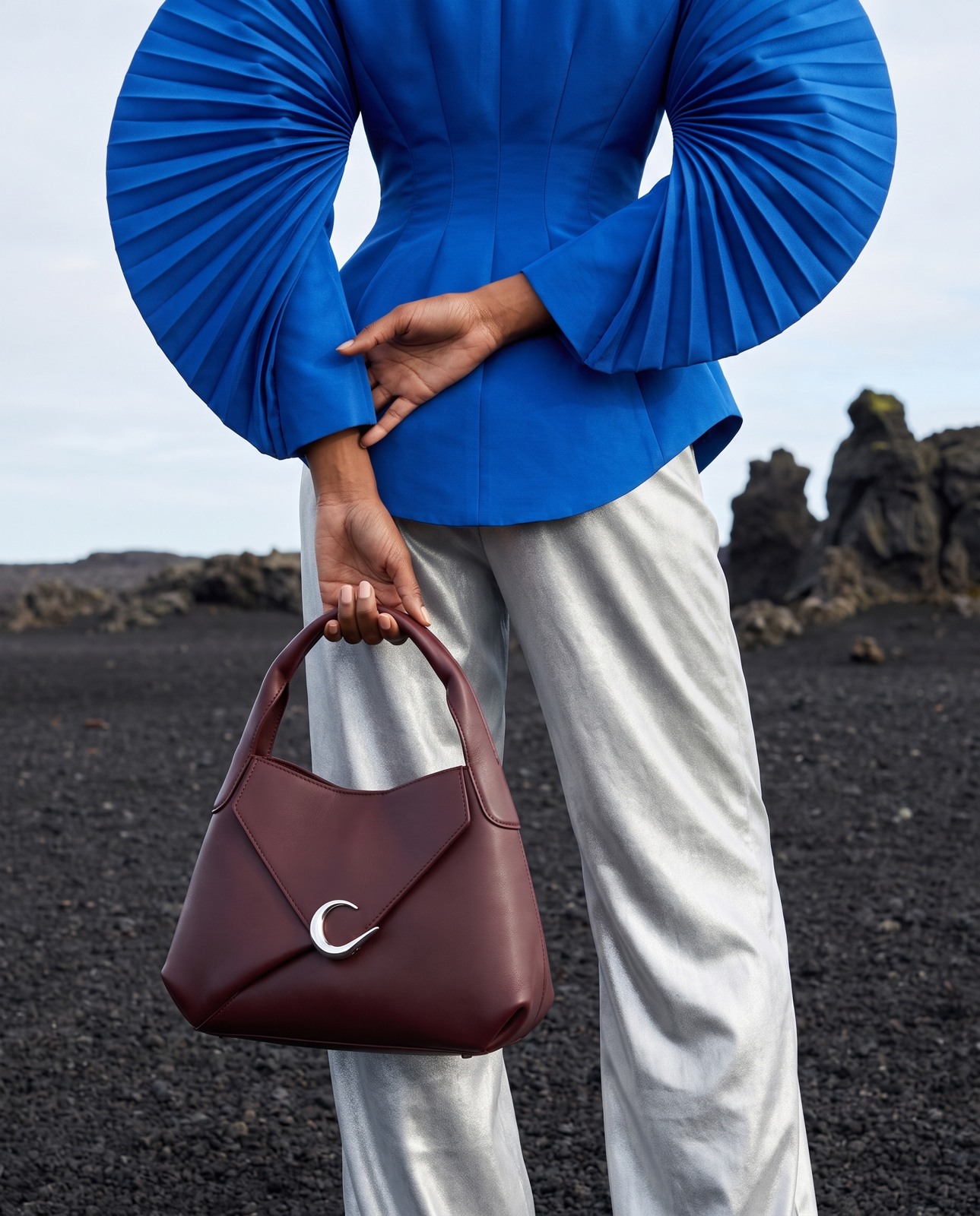 AI fashion campaign: cobalt-blue pleats, silver trousers and a burgundy handbag against volcanic rock